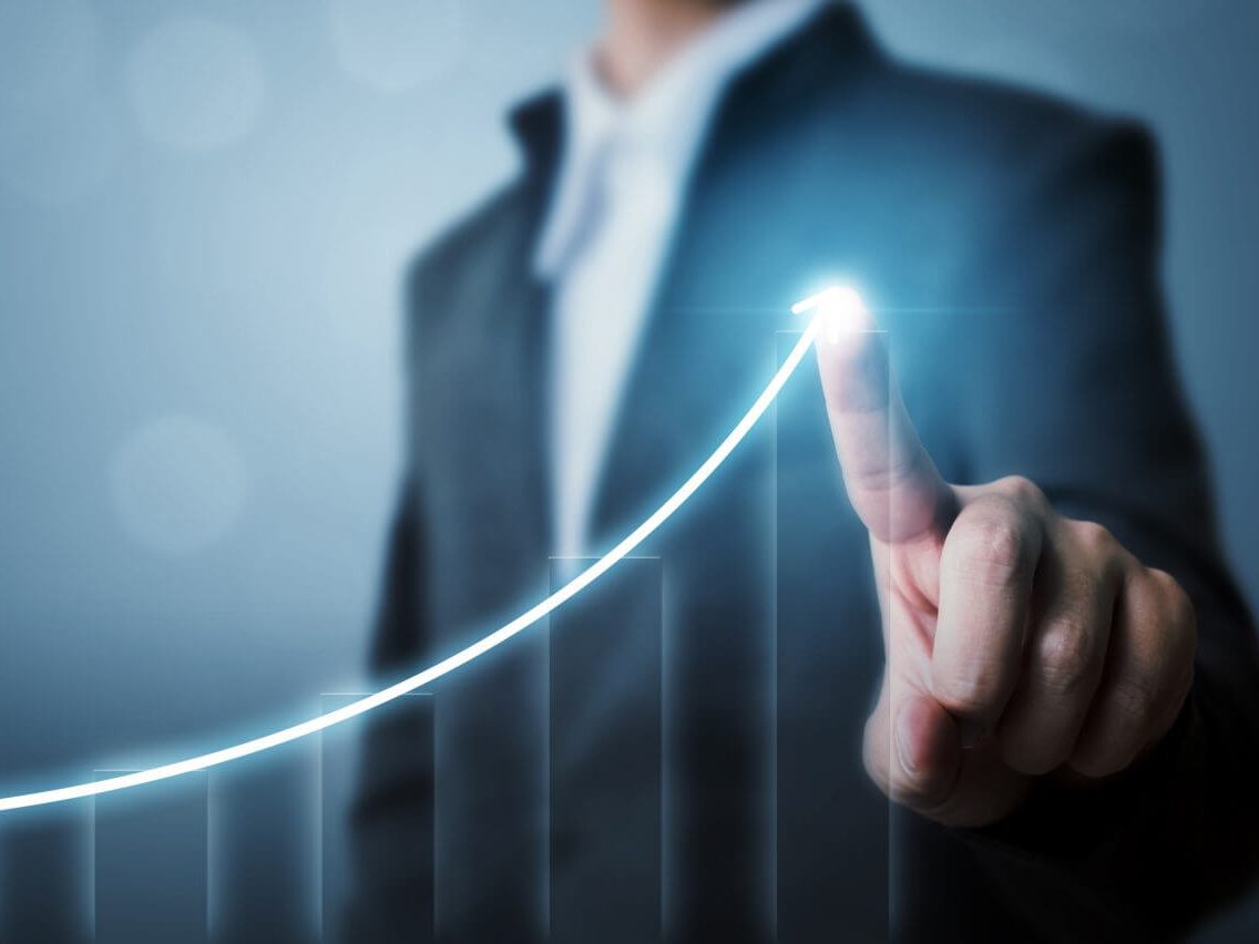 A man is pointing at the screen where an ascending bar graph glows with a brighter glowing line showing the ascension and ending at his finger. 

"Accelerate Success" and "Maximize Results" are two benefits of business coaching.