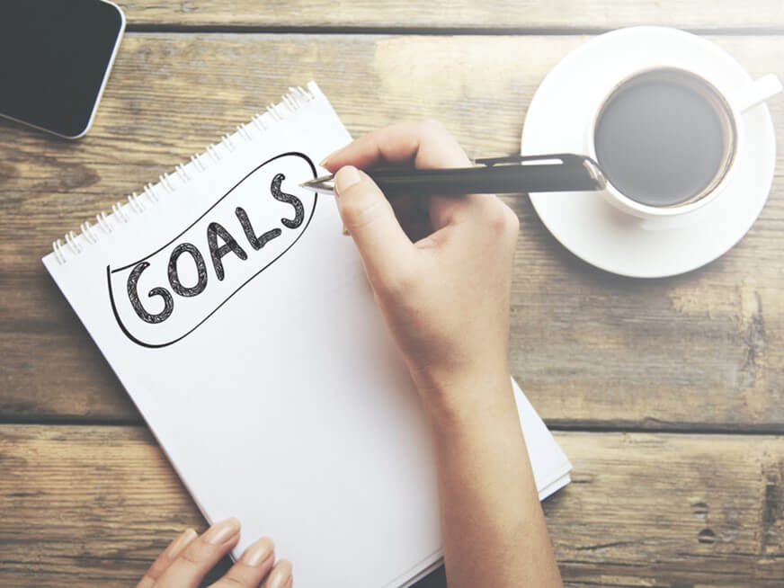 A notebook with the text "GOALS" written across the top on a table with a coffee mug and the edge of a screen.
Beginning Goal Setting