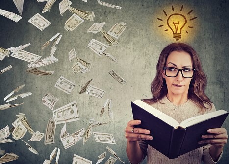A woman hold an open book in front of her. Above her head is a lit up lightbulb. She is looking to the side where money is falling like rain.