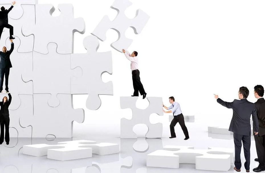 Two people in business attire are acts as managers to a group of other people in business attire as they move white puzzle pieces as tall as they are into place with the puzzle stand straight up. Three people are standing on top of each other so that the top person can reach a puzzle piece high up. The other two people are also helping each other, with the one on the ground pushing a puzzle piece into place with the other standing on top of that piece placing his piece in a higher position. (ie. example of team work and management)