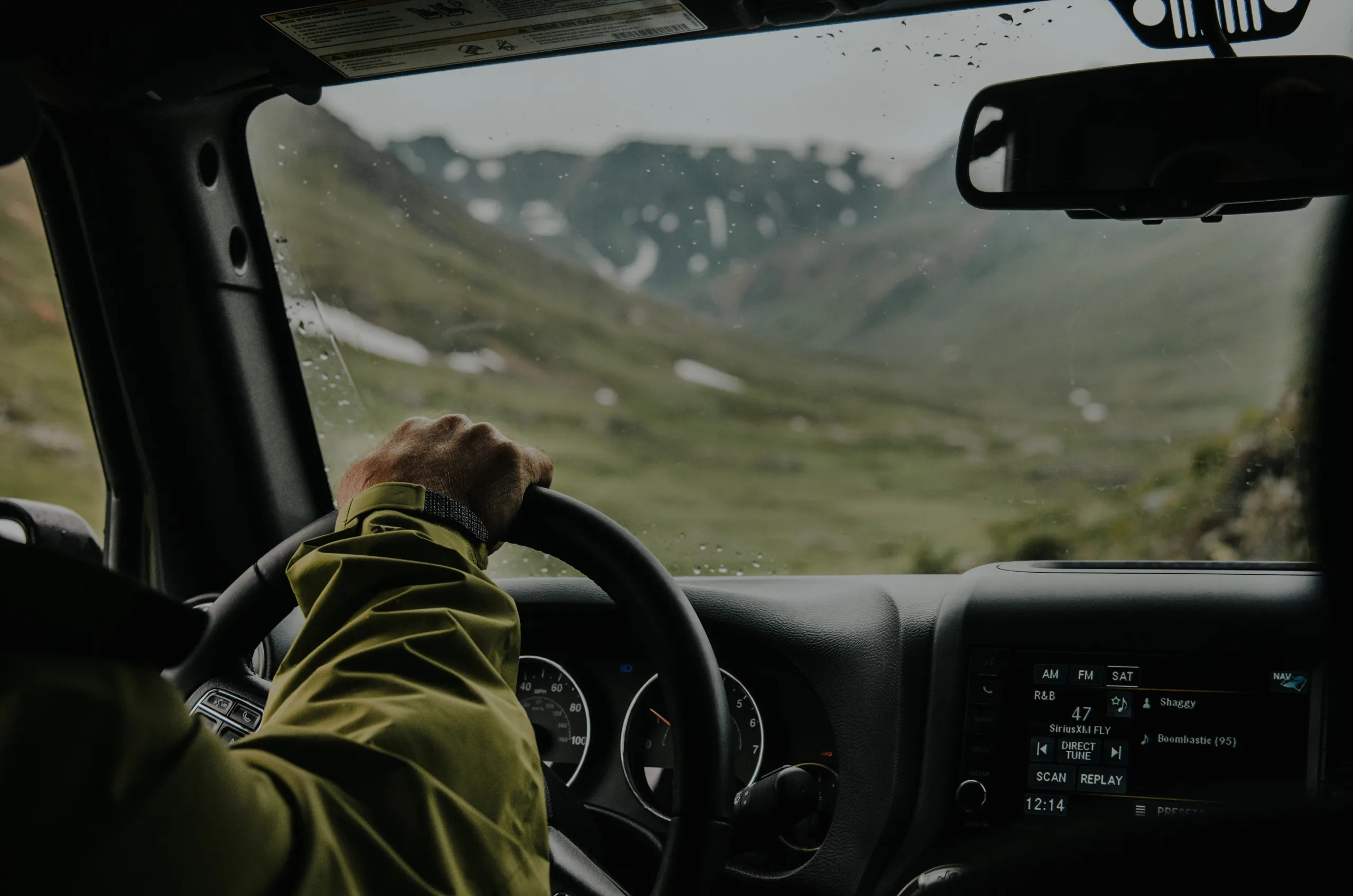 View of a rainy day in mountains through the windshield of a car. A hand and arm are steering the car.
(Aligned and Primed)