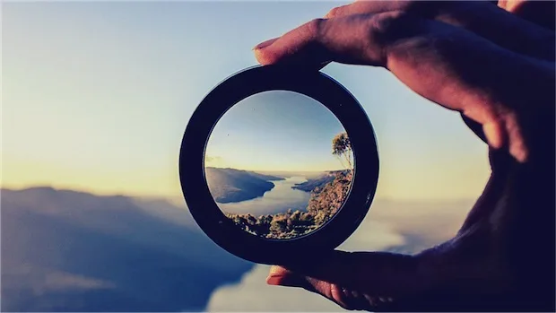 A hand holds a magnifying lenses that brings a landscape image into focus.